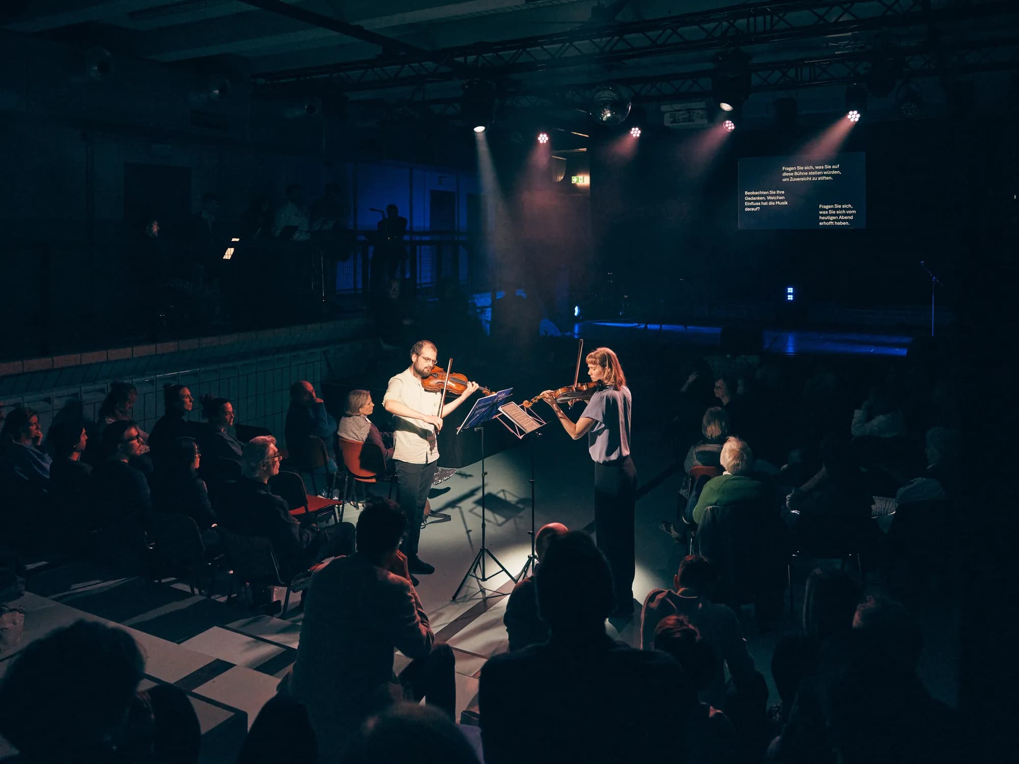 Duo Zwischen im Auftritt — Annika Spegg und Robert Rülke spielen Geige im Spotlight, im Hintergrund die Projektion „Fragen Sie sich, was Sie auf diese Bühne stellen würden, um Zuversicht zu stiften.“